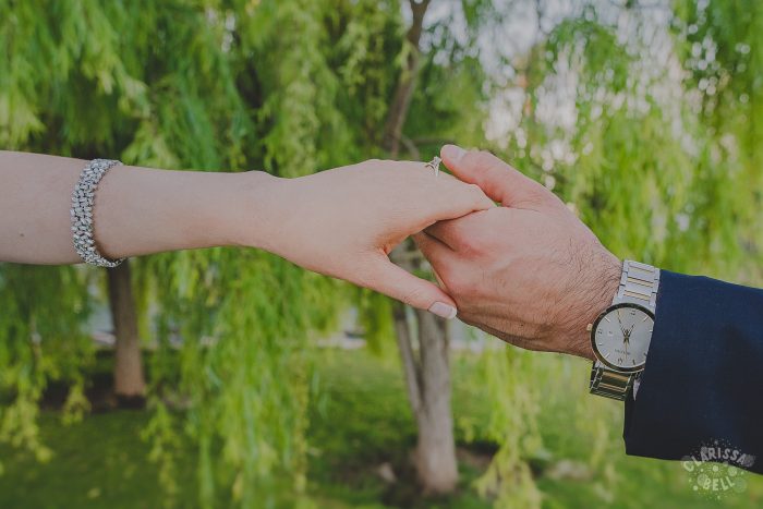 Novio tomando delicadamente la mano de su novia en la sesión de fotos de su boda, al fondo hay árboles verdes y contrasta mucho las manos deel fondo